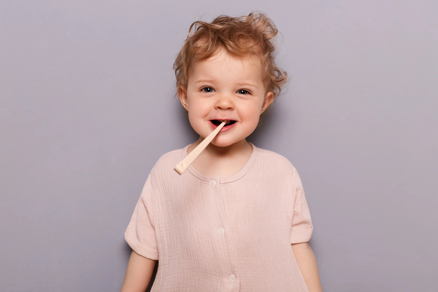 Por que usar pasta de dente sem flúor ao escovar os dentes das crianças? Criança sorridente com cabelo cacheado e uma escova de bambu na boca. A imagem mostra alegria e descontração em um fundo cinza.
