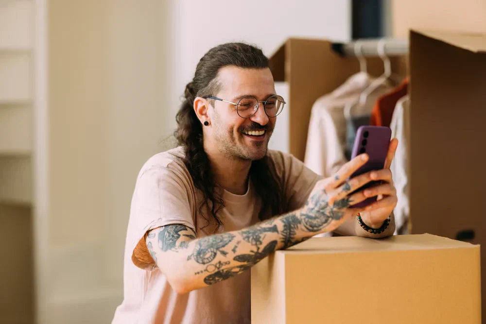 Cuidados com cabelo longo masculino: principais dicas pra ter fios sempre saudáveis Homem sorridente usando celular ao lado de caixas de mudança, com tatuagens e cabelo comprido em ambiente iluminado.