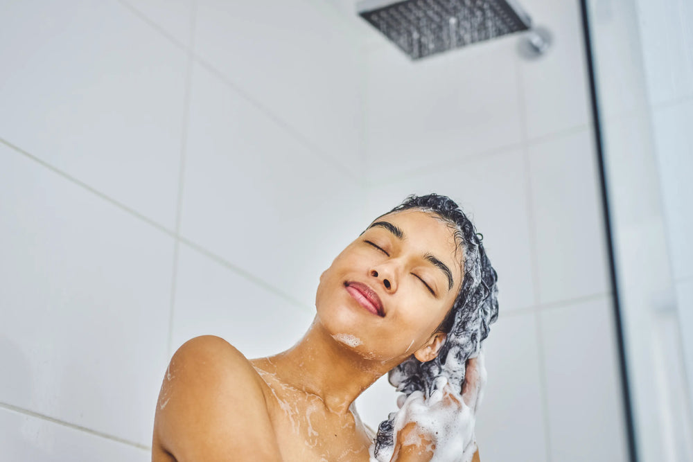 Descubra o Low Poo: as melhores técnicas e produtos pra investir Mulher sorrindo enquanto aplica shampoo no cabelo no chuveiro, evidenciando cuidados com os cabelos e bem-estar durante o banho.