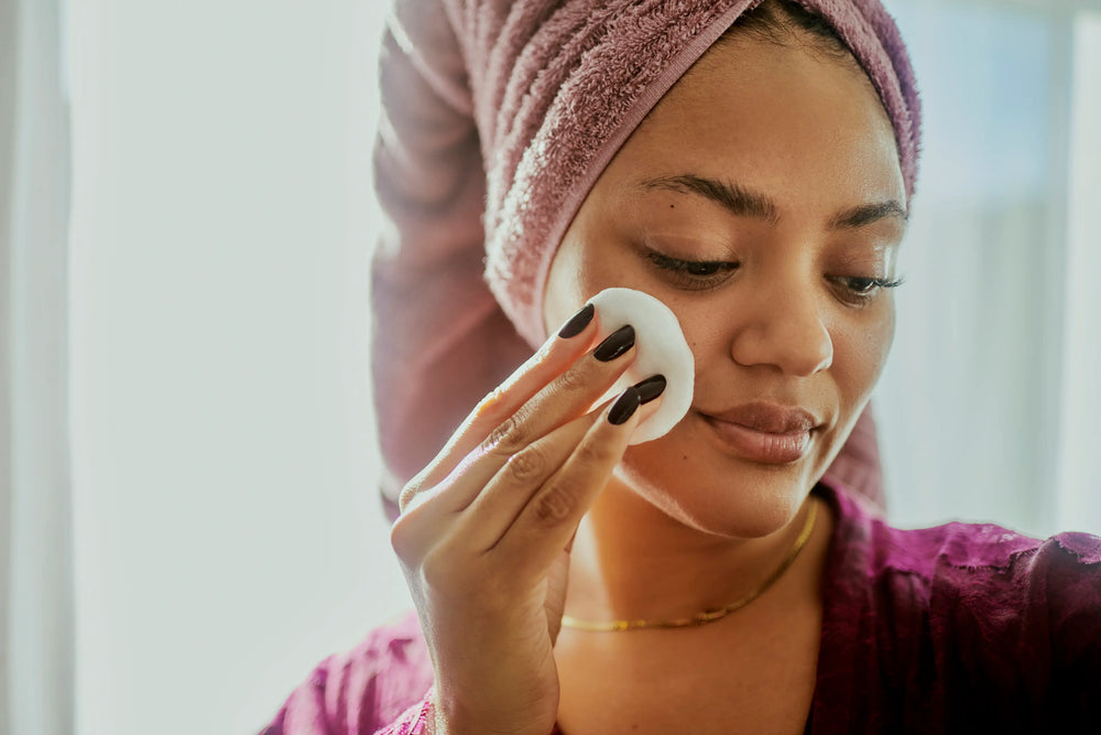 Como fazer limpeza de pele em casa? 4 dicas práticas! Mulher com toalha na cabeça fazendo limpeza facial com disco de algodão, destacando cuidados com a pele e beleza.