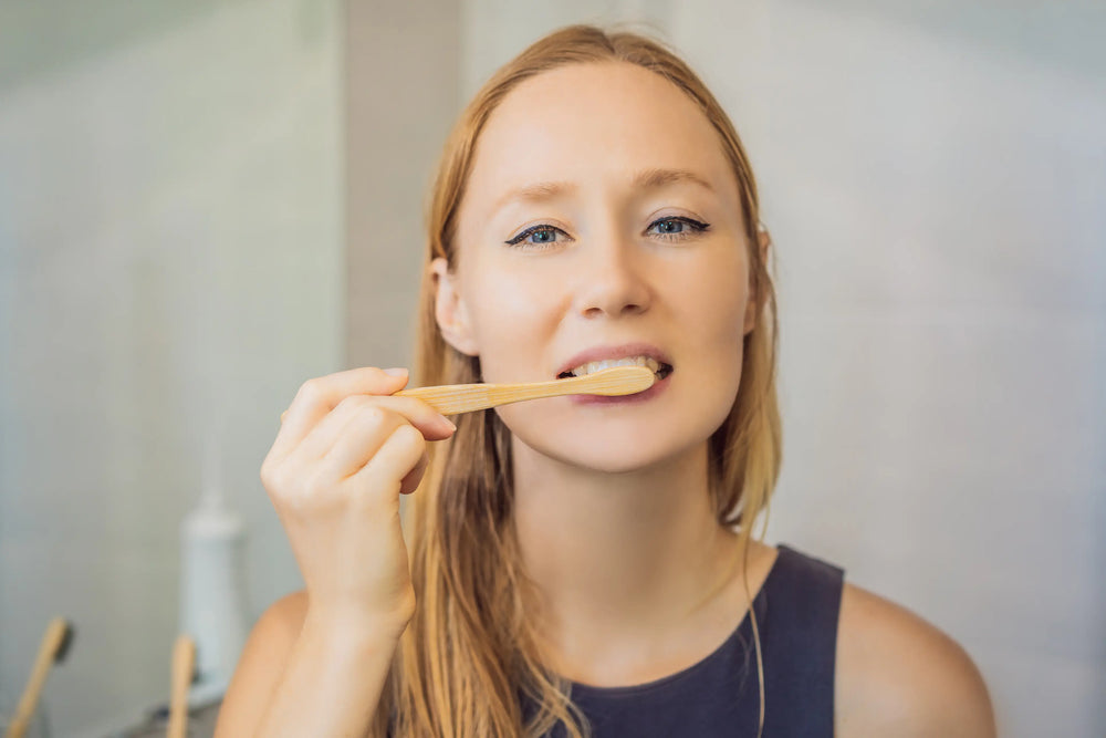 Posso usar pasta de dente com flúor todos os dias? Uma mulher sorridente escovando os dentes com uma escova de bambu em um ambiente moderno e aconchegante, promovendo hábitos saudáveis de higiene bucal.