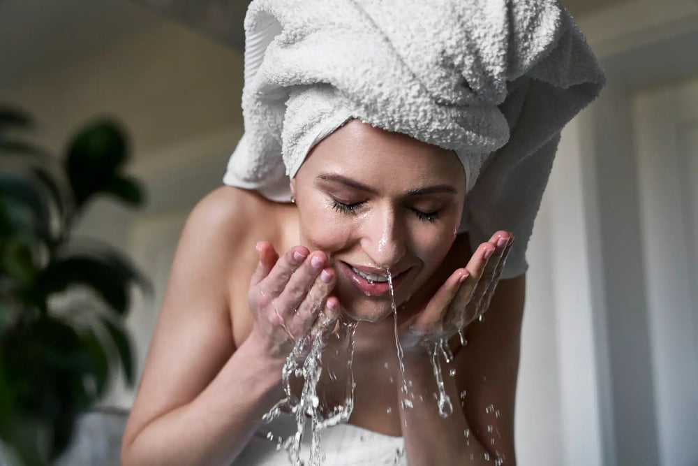 Skincare noturno: saiba a ordem, melhores produtos e dicas essenciais Mulher lavando o rosto com água em um ambiente iluminado, com toalha na cabeça, destacando cuidados pessoais e beleza.