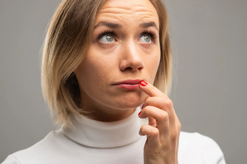 Afta: o que é, como surge e como evitar? Mulher pensando, olhando para cima com expressão contemplativa, usando blusa branca e destacando unhas vermelhas.