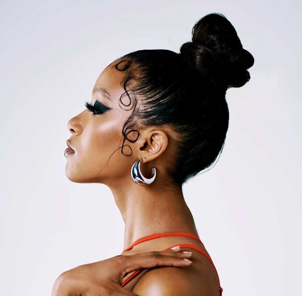 Baby hair: entenda o que é, como fazer, controlar e fortalecer os fios Imagem de perfil de uma mulher com cabelo preso em um coque alto, usando brincos prateados e uma blusa vermelha, destacando um olhar elegante.
