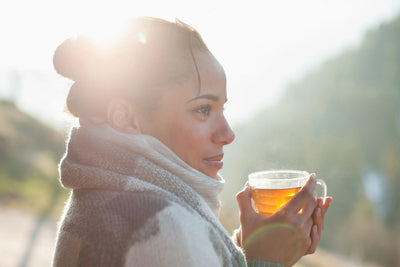 Mulher segurando uma caneca de chá, contemplando a paisagem iluminada pelo sol. A imagem transmite tranquilidade e conexão com a natureza.