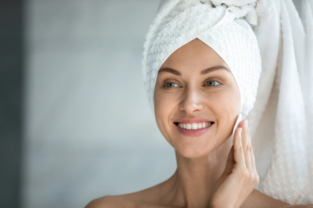 Como tirar maquiagem sem demaquilante? Mulher sorridente com toalha na cabeça, cuidando da pele em ambiente claro.