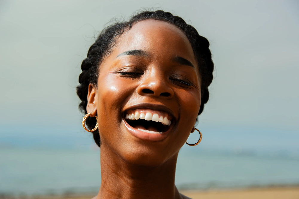 Qual o melhor fator de protetor solar para pele negra? Mulher sorrindo com alegria em um cenário ao ar livre, destacando seu sorriso largo e feliz, refletindo um momento de felicidade.