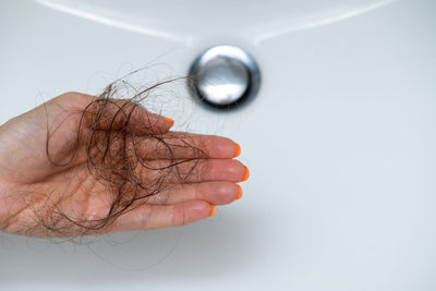 Mão segurando fios de cabelo soltos, evidenciando a questão da queda de cabelo e cuidados capilares em um ambiente de banheiro.