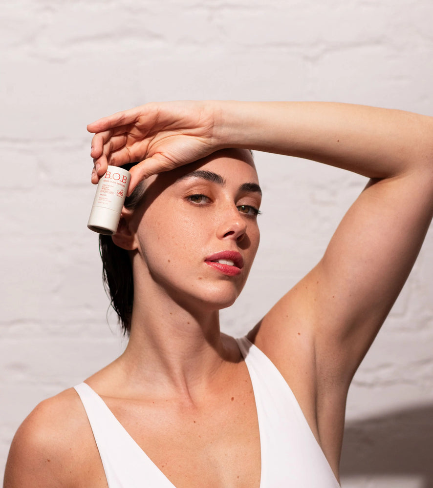 Modelo segurando um frasco de produto B.O.B. próximo ao rosto, com fundo neutro e expressão confiante.