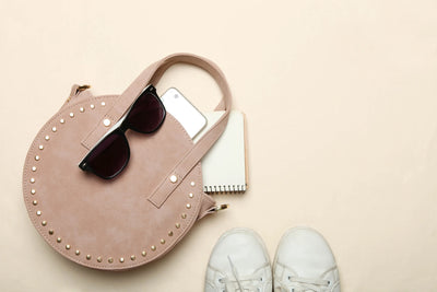 Imagem de uma bolsa redonda rosa com detalhes em dourado, óculos escuros, um celular e um caderno ao lado de tênis brancos.