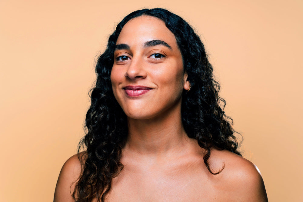 Retrato de uma mulher com cabelo cacheado e pele clara, sorrindo em frente a um fundo laranja. A imagem transmite uma sensação de alegria e confiança.