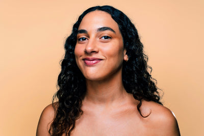 Retrato de uma mulher com cabelo cacheado e pele clara, sorrindo em frente a um fundo laranja. A imagem transmite uma sensação de alegria e confiança.