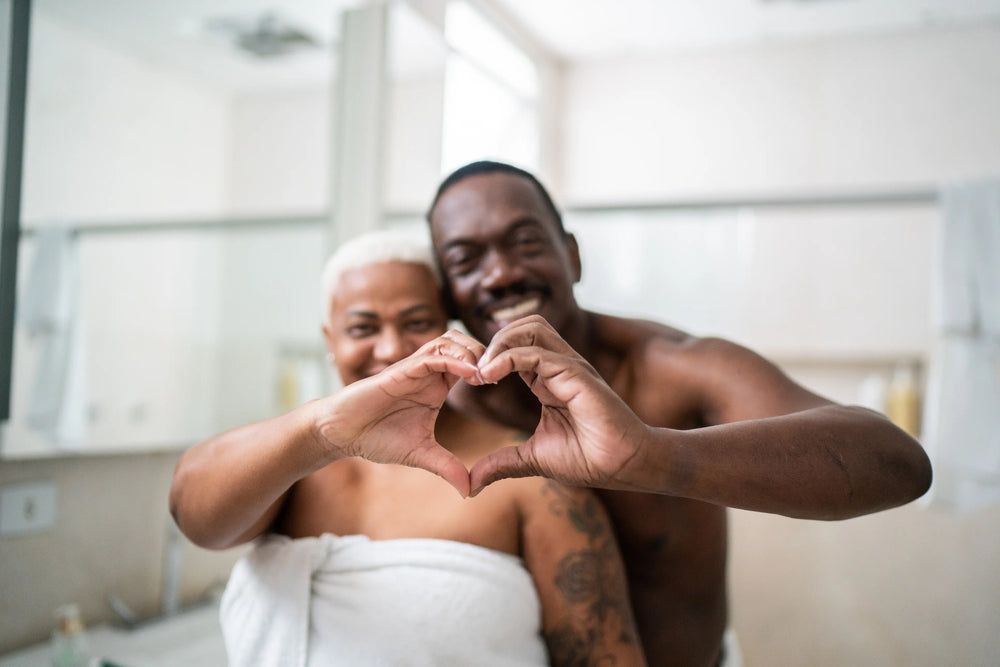 Casal feliz mostrando coração com as mãos no banheiro, em um ambiente acolhedor.