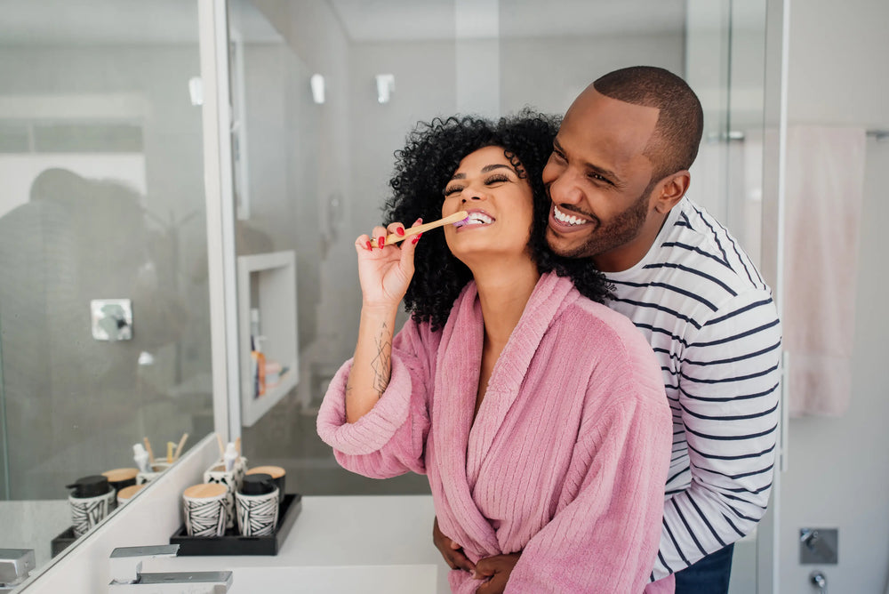 Casal sorridente em banheiro, ela escovando os dentes e ele a abraçando. Um momento de alegria e cuidado pessoal juntos.