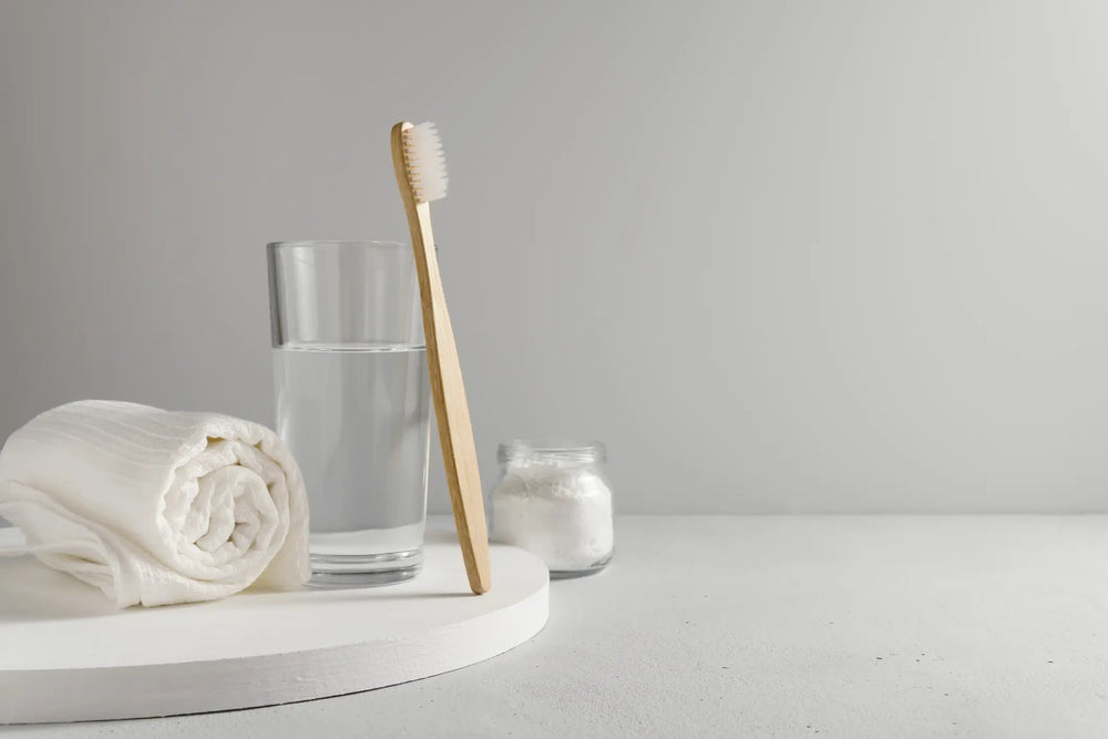 Imagem que ilustra itens de higiene pessoal, com uma escova de dentes de bambu, um copo de água e uma toalha branca.
