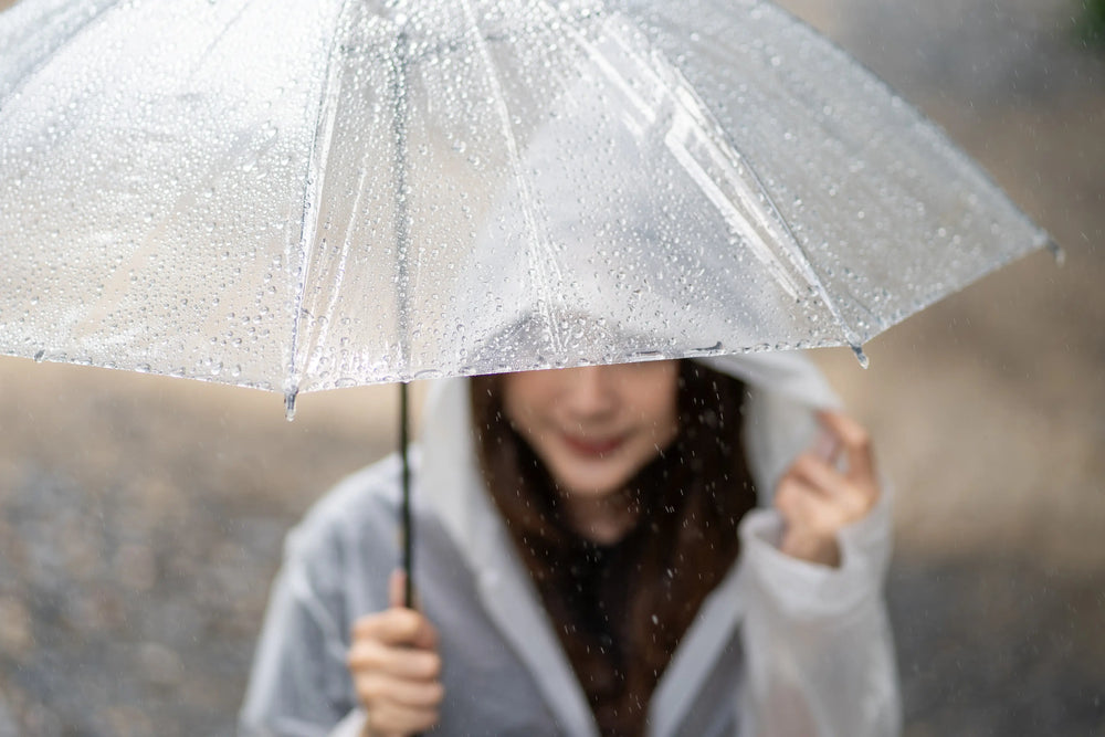 Imagem de uma jovem segurando um guarda-chuva transparente em um dia chuvoso. A cena captura gotas de chuva e transmite um sentimento de introspecção em um ambiente natural.