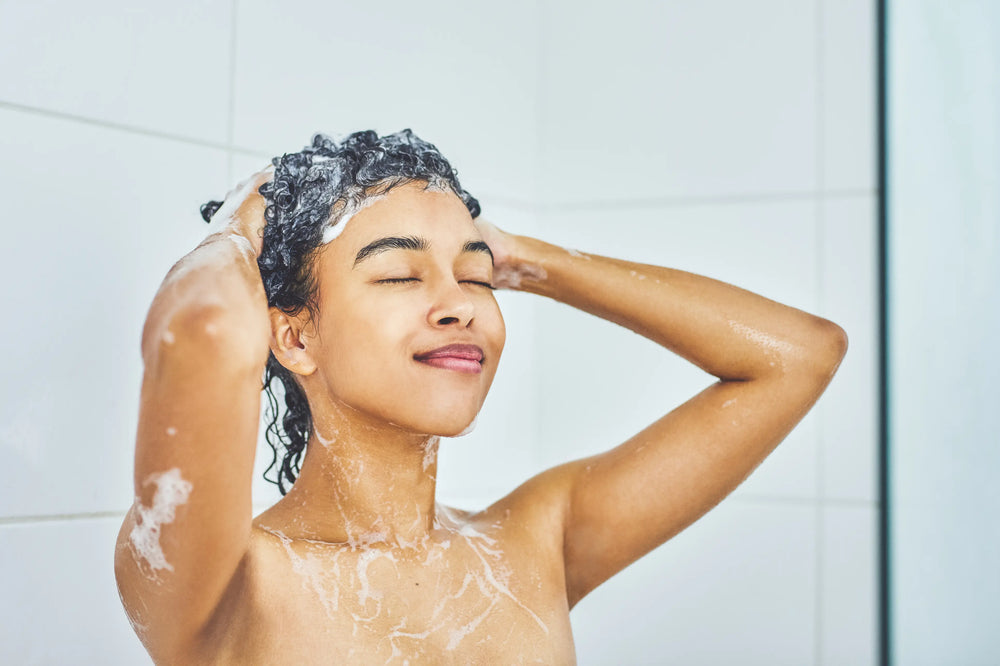 Mulher sorridente lavando o cabelo no chuveiro, com espuma de shampoo e expressão de relaxamento, representando a rotina de cuidados pessoais.