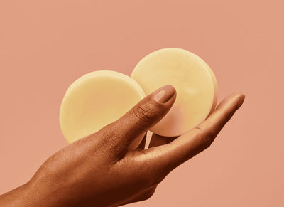 Mão segurando dois condicionadores em barra redondos em um fundo suave, destacando a estética e a beleza dos produtos de cuidados pessoais.