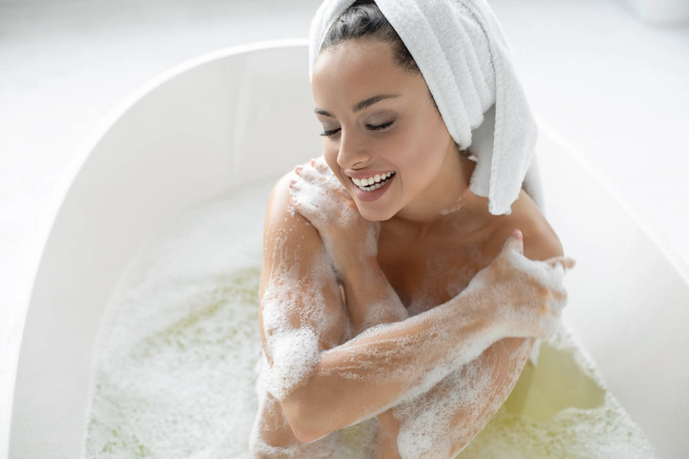 Mulher sorrindo enquanto toma banho em uma banheira branca, cercada de espuma relaxante.