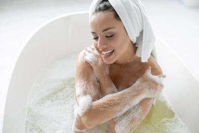 Mulher sorrindo enquanto toma banho em uma banheira branca, cercada de espuma relaxante.