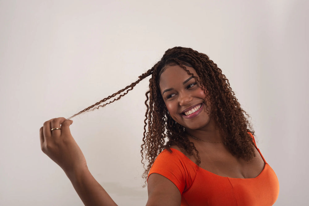 Mulher sorridente com cabelo cacheado, usando blusa laranja, segurando uma mecha de cabelo de forma descontraída. A imagem transmite alegria e estilo.