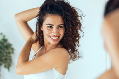 Mulher sorridente com cabelo cacheado olhando no espelho, destacando a beleza dos cabelos.