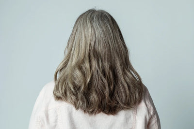 Imagem de uma mulher com cabelo longo e ondulado, visto de costas. Os cabelos são grisalhos, com um tom natural e saudável. A cena é minimalista, com fundo claro.