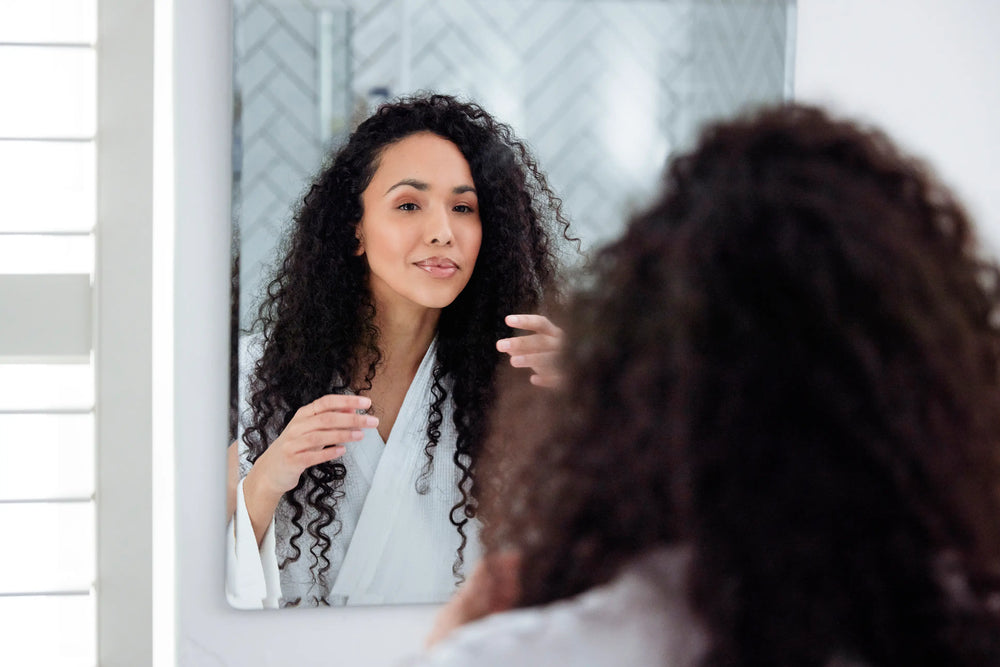Mulher de cabelos cacheados se arrumando em frente ao espelho, usando roupão branco e com um sorriso no rosto, refletindo cuidado com a beleza em um ambiente iluminado.