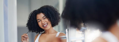 Mulher sorridente com cabelo cacheado se olhando no espelho, refletindo autoestima e beleza natural.