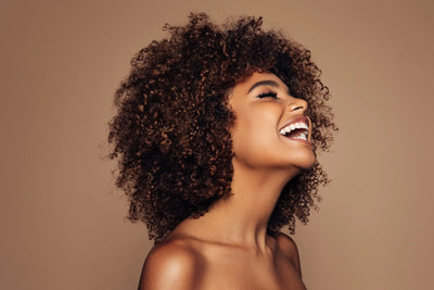 Uma mulher com cabelos cacheados e volumosos sorri amplamente, destacando sua beleza natural e radiante. O fundo neutro ressalta seu sorriso e a textura do cabelo.