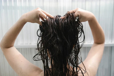 Mulher com cabelos longos e molhados, segurando os fios na parte de trás da cabeça, em ambiente claro e moderno.