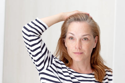 Mulher com blusa listrada, expressão serena e olhar natural. Cabelo solto e levemente ondulado. Fundo neutro que destaca a suavidade de seu rosto.