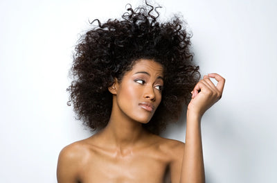 Mulher com cabelo cacheado volumoso e expressão pensativa, destacando sua beleza natural em fundo neutro.