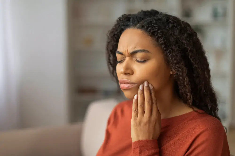 Mulher com expressão de dor facial, tocando a mandíbula em um ambiente interno. A imagem retrata o desconforto e a preocupação com a saúde bucal.