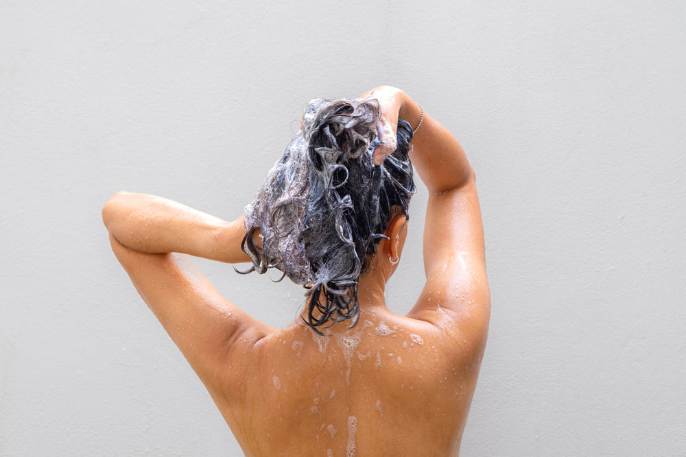 Mulher lavando o cabelo com shampoo. Os fios estão ensaboados, mostrando cuidado com a higiene capilar. A imagem destaca a importância de uma rotina de cuidados com os cabelos.