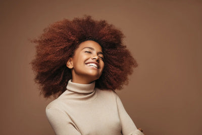 Mulher sorridente com cabelo crespo volumoso, usando suéter de gola alta em fundo neutro, destacando beleza natural e autoconfiança.