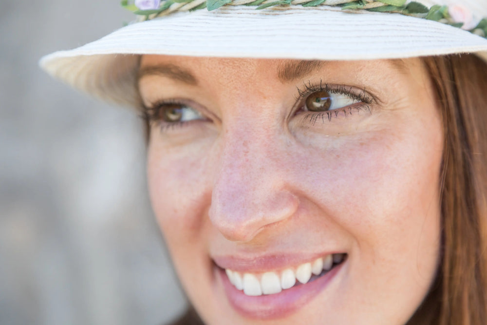 Mulher sorridente com chapéu e flores, mostrando atitude alegre em um dia ensolarado.