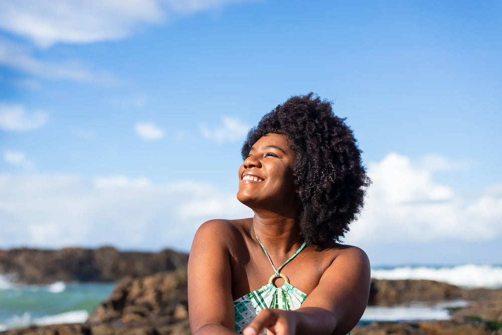 Mulher sorridente com cabelo crespo, usando roupa de verão, desfrutando de um dia ensolarado na praia com céu azul atrás.