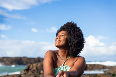 Mulher sorridente com cabelo crespo, usando roupa de verão, desfrutando de um dia ensolarado na praia com céu azul atrás.