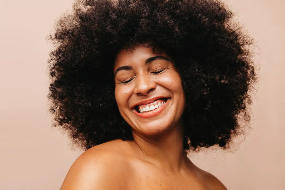 Mulher sorrindo com cabelo cacheado volumoso em fundo neutro, mostrando felicidade e confiança. Ideal para temas de beleza e cuidados capilares.