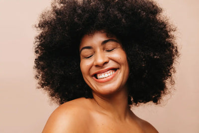 Mulher sorrindo com cabelo cacheado volumoso em fundo neutro, mostrando felicidade e confiança. Ideal para temas de beleza e cuidados capilares.