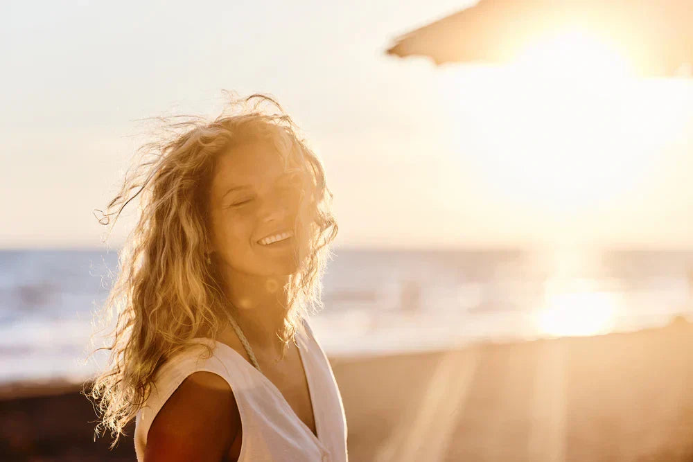Mulher sorrindo na praia ao pôr do sol, com cabelos ondulados e iluminada pela luz dourada do sol. A cena transmite alegria e tranquilidade em um ambiente natural.