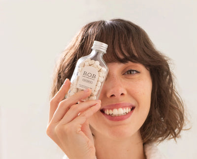 Mulher sorrindo e segurando um frasco de tabs dentais B.O.B. com pastilhas brancas, destacando a boa saúde e beleza.