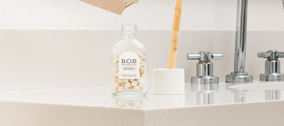 Imagem de produtos B.O.B. para cuidados dentais, incluindo frasco com pastilhas e escova de dentes, sobre bancada de banheiro moderna.