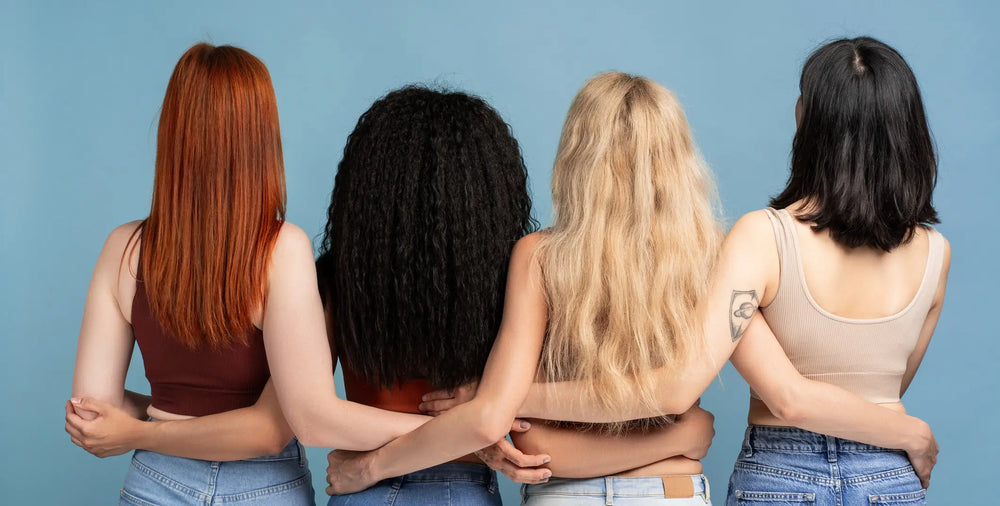 Quatro mulheres de diferentes estilos de cabelo estão abraçadas, vista de costas, em um fundo azul. Elas usam roupas casuais e jeans, representando a diversidade feminina.