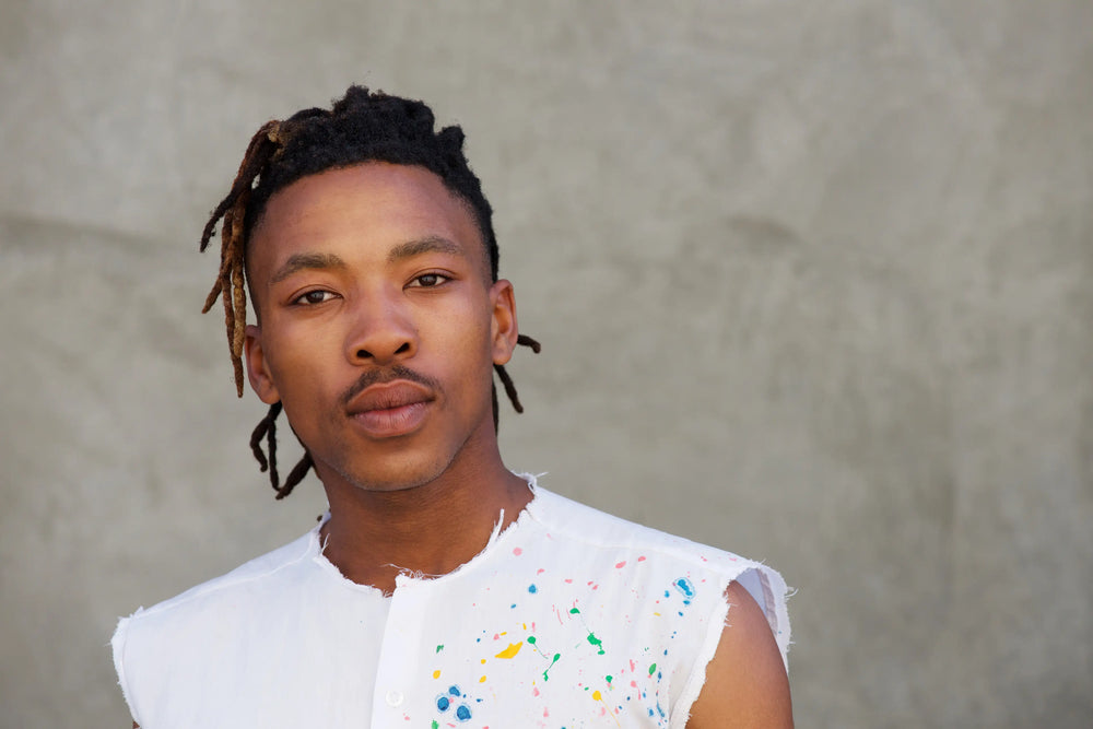 Retrato de jovem homem com cabelo com dreads e camisa branca. Ele apresenta uma expressão confiante, posando em fundo neutro.
