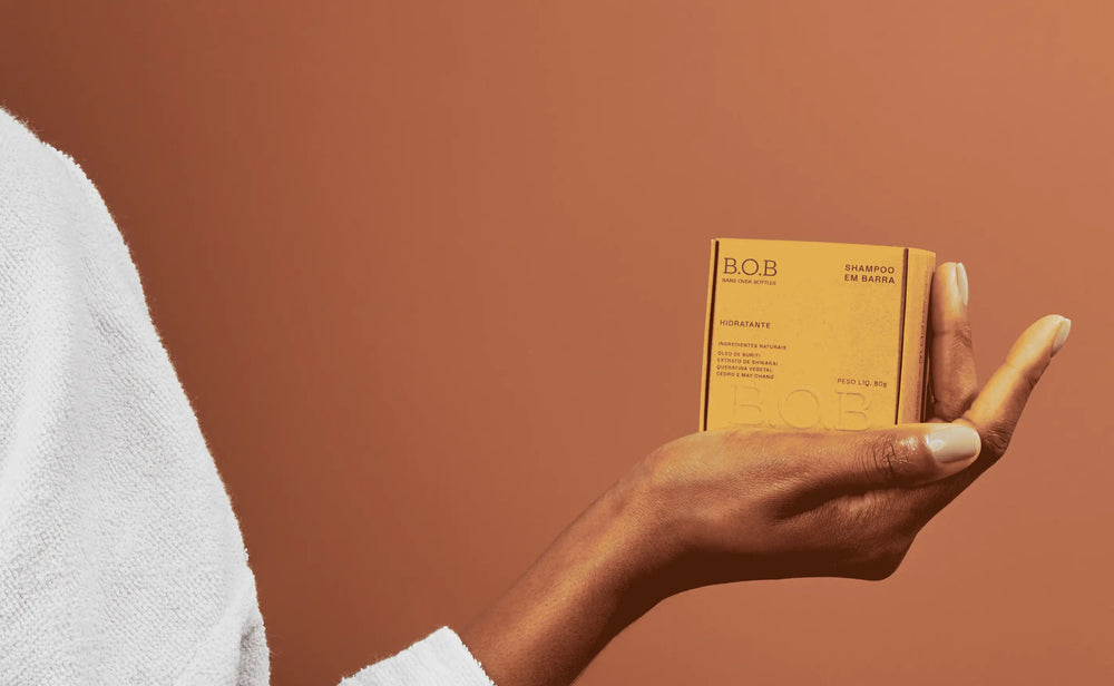 Imagem de uma pessoa segurando um shampoo em barra da marca B.O.B, em embalagem amarela, contra um fundo terroso, ideal para cuidados capilares.